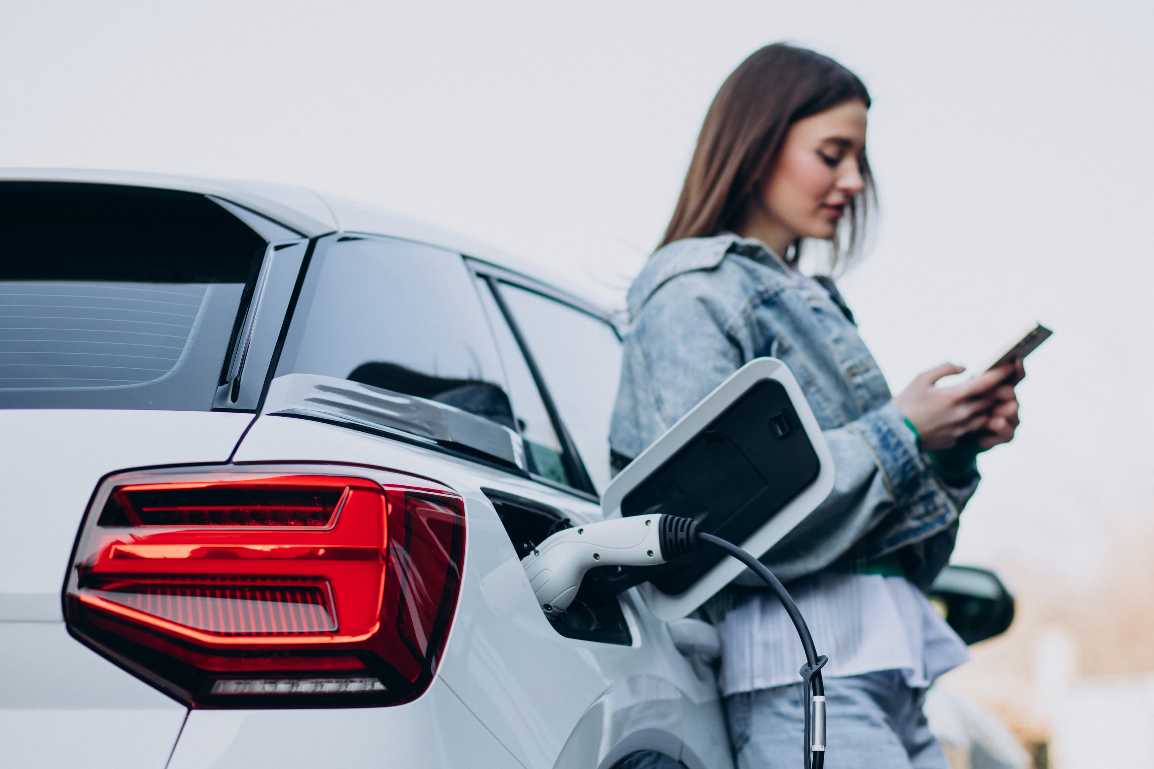 Mujer junto a su coche eléctrico blanco conectado a un cargador, consultando el móvil